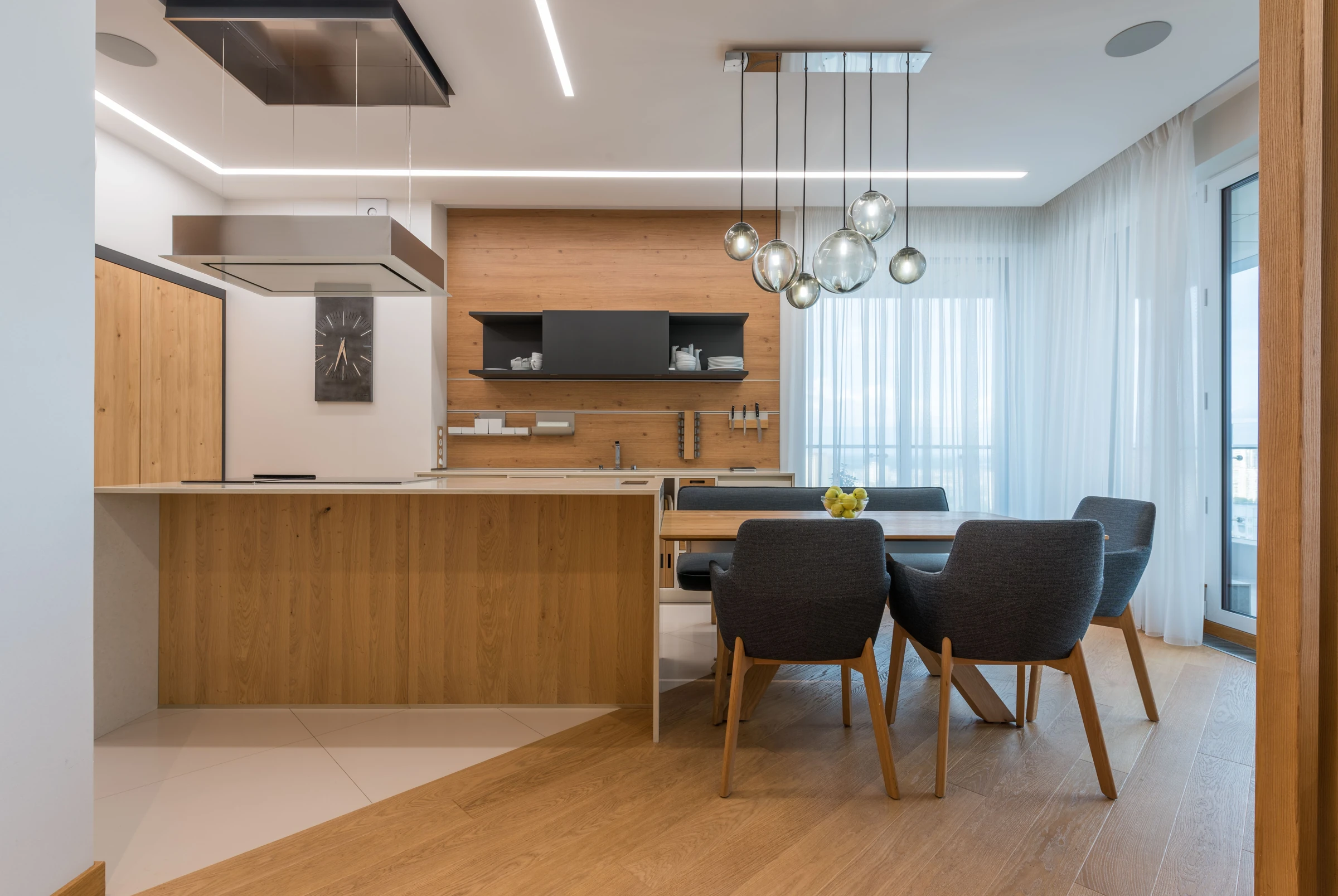 Warm wood kitchen with pale counters, upholstered grey dining chairs and suspended glass globe lights.